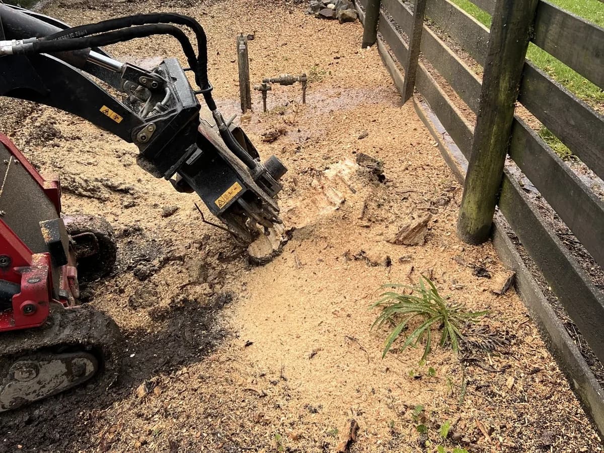 A stump grinder attachment cutting into a stump, with sawdust spread across the ground beside a timber post-and-rail fence