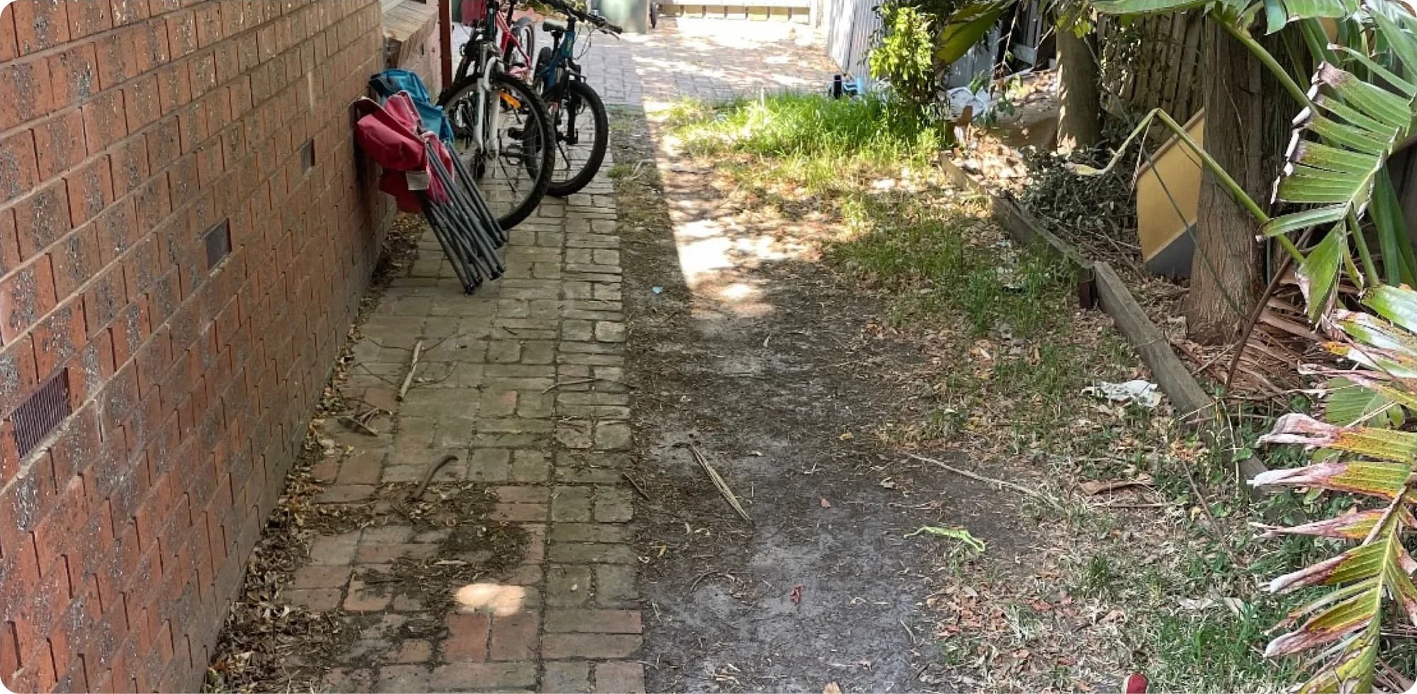 Narrow side path of uneven pavers beside a brick wall, littered with debris